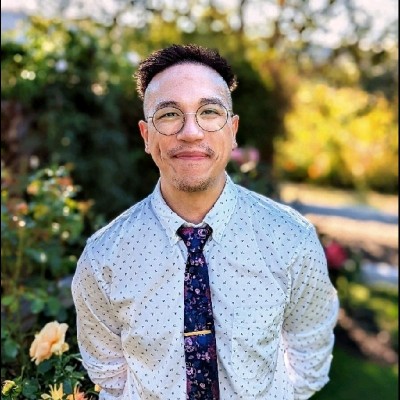 Josh Baez, senior manager of demand generation for Netline, smiles in a white shirt and dark print tie in front of green leafy trees. 