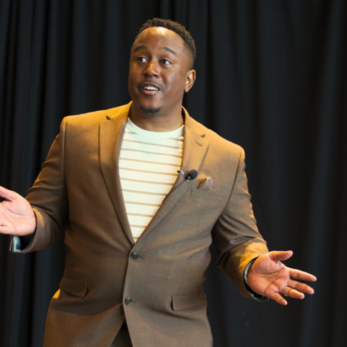 Troy Sandidge, founder of Strategy Hackers, gestures while wearing a brown jacket in front of a black background. 