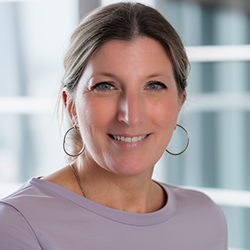 Andi Robinson, content strategist with Hijinx Media, smiles in a purple top and hoop earrings in front of white-paned windows. 