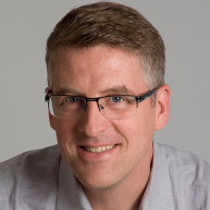 Tony Byrne, founder of Real Story Group, smiles in a grey shirt and dark-framed glasses in front of a grey background. 