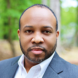 A. Lee. Judge, founder and CMO of Content Monsta, smiles in a grey suit coat and white shirt in front of green leafy trees.