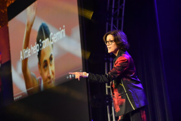 Ann Handley is wearing black glasses and a metallic burgundy suit pointing at the audience. The slide in the background says "A little help from Gemini."