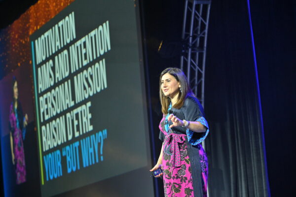 Shabnam Mogharabi is standing on stage wearing a navy and pink floral dress. The slide in the background says "Motivation aims and intention personal mission raison d'etre your 'But Why?'"