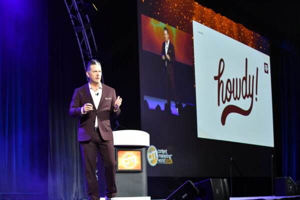 R. Ethan Braden standing on stage in a black suite and white button down shirt. The slide in the background says "howdy!"