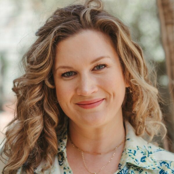 Allison Champion has curly blonde hair and is wearing a cream and blue floral blouse.