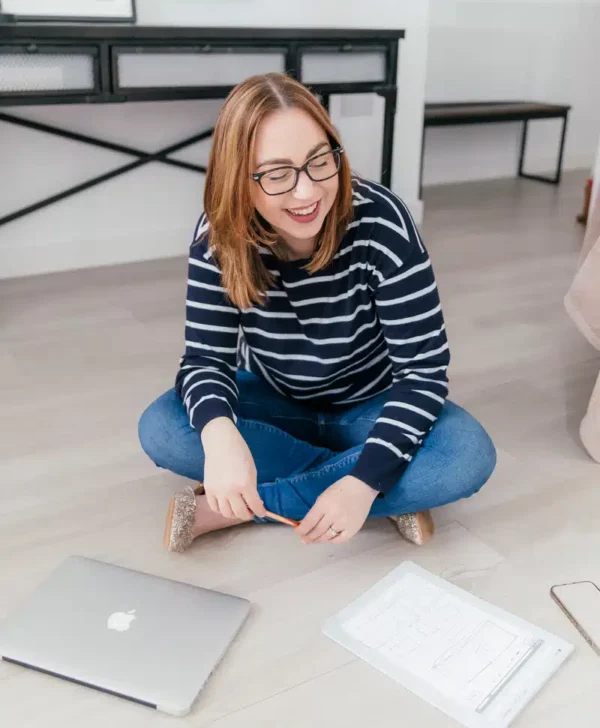 Kate is sitting on the floor wearing a blue and white sweater and blue jeans. She is wearing glasses, looking down smiling.